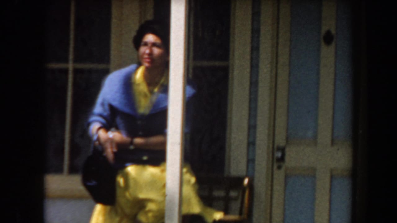 In 1961 California home-movie footage, a woman in a blue jacket and yellow skirt poses near glass-paneled doors while holding a black handbag.