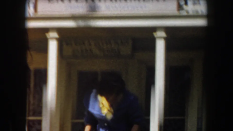 The woman leans forward in the shaded entrance area, with porch columns and a partially visible sign above her.