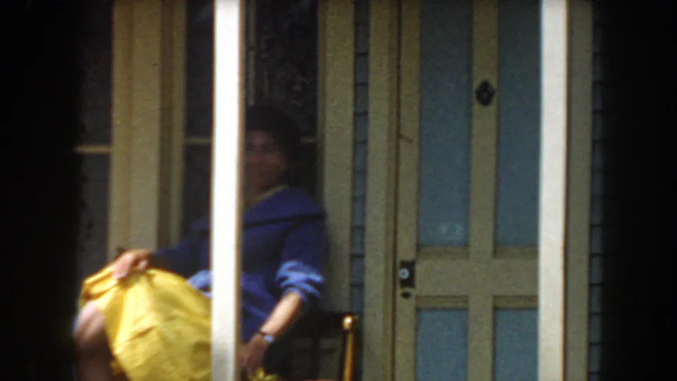 The woman appears seated or leaning near the glass-paneled doorway, partly hidden by a porch column, with her yellow skirt prominent.