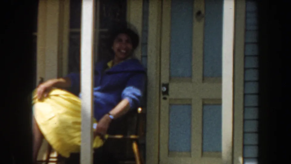 1961 California home-movie frame of a smiling woman in a blue jacket and yellow skirt seated on a porch chair.