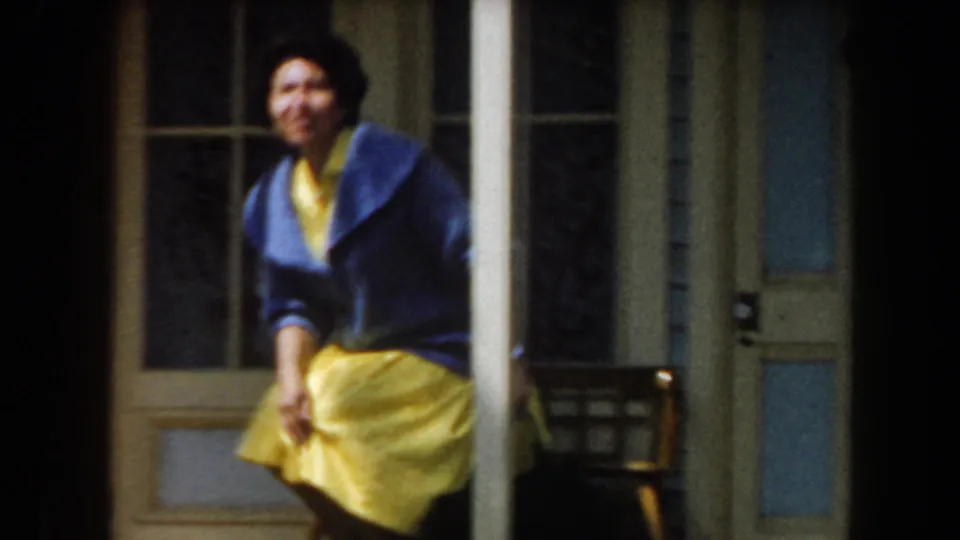 1961 California frame showing the woman seated in front of the storefront, with columns, windows, and a nearby chair visible.
