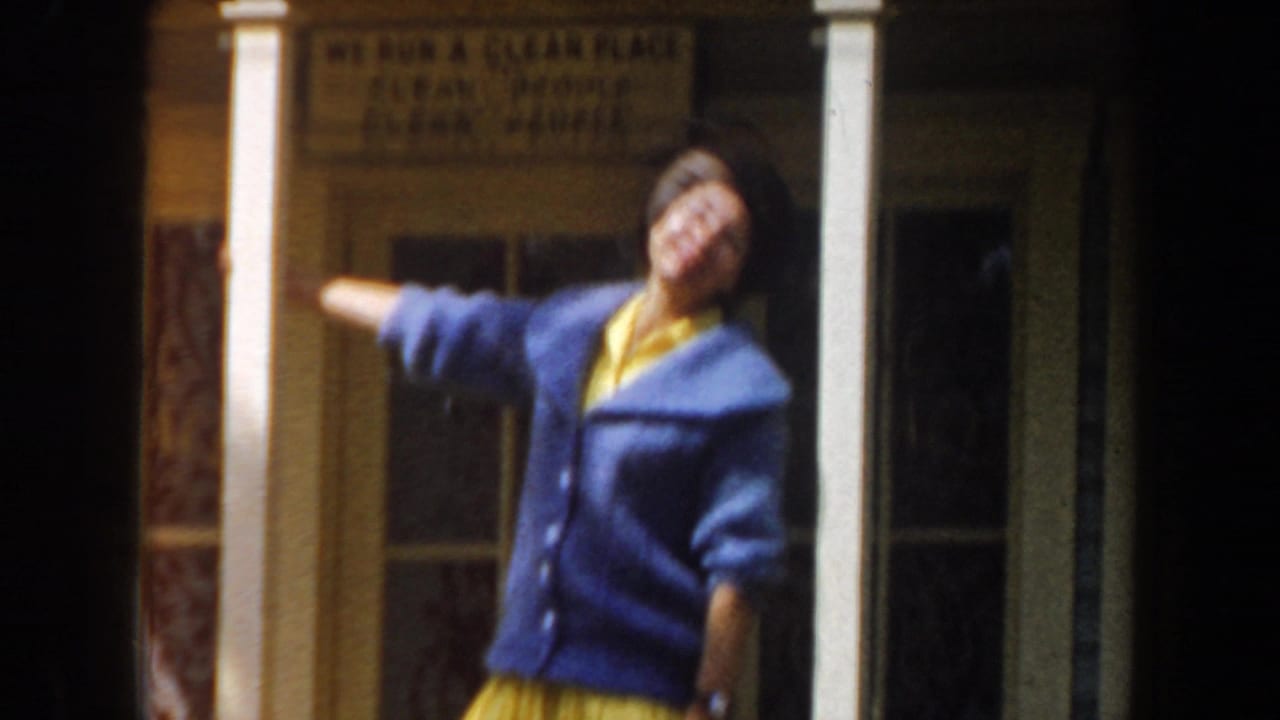Young woman in a blue cardigan dances near a porch with a sign behind her.