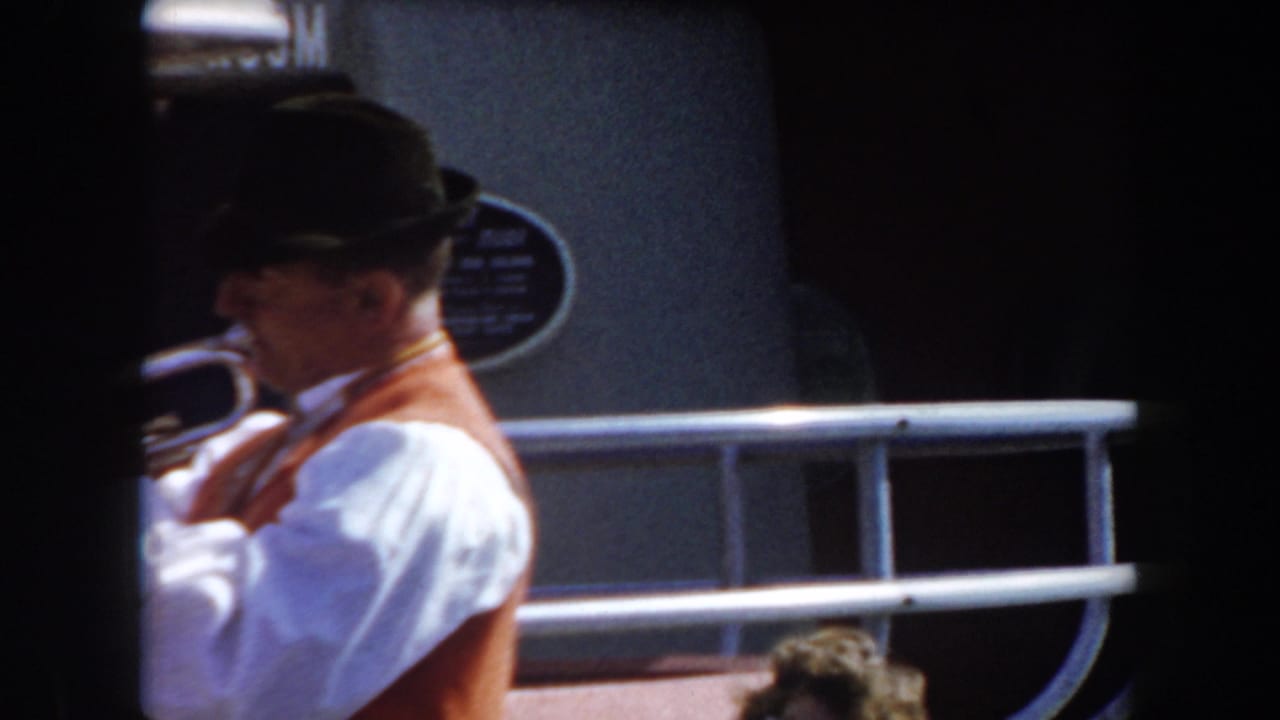 A brass musician in a dark hat plays beside a railing and building wall.