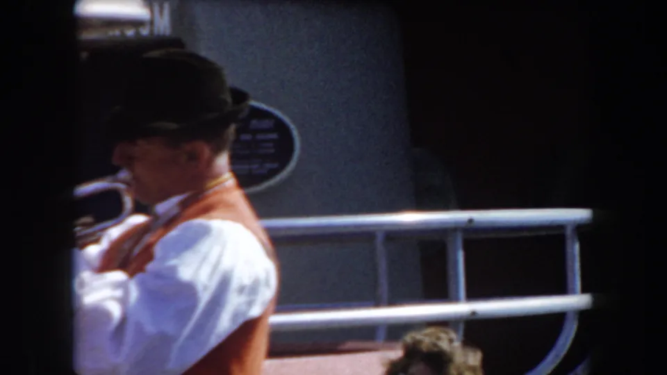 A brass-band musician in a dark hat and orange vest plays near a railing in the 1961 California street scene.