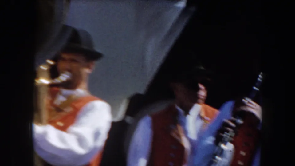 Two band members in dark hats and orange vests perform in a soft, grainy 1961 California home-movie frame.
