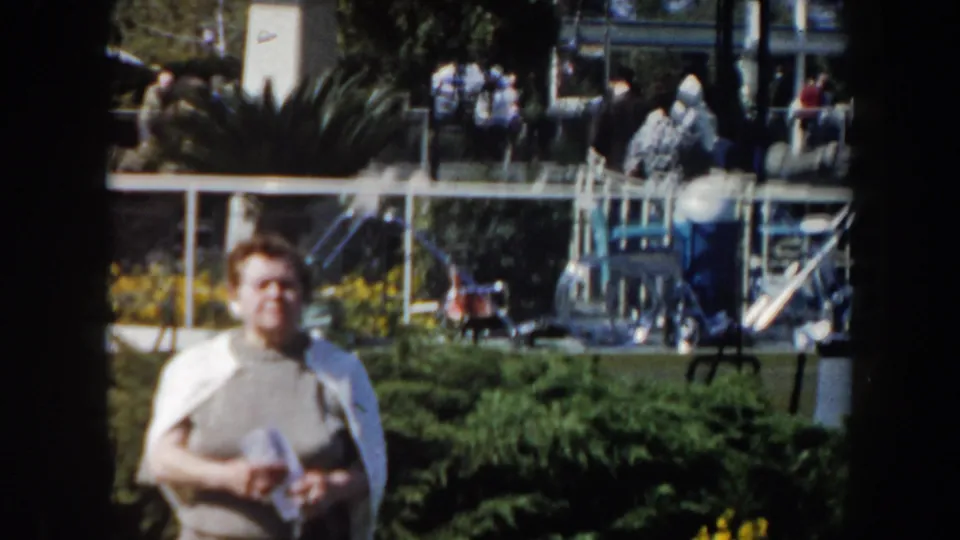 1961 California home-movie frame showing a woman in the foreground near landscaping, railings, and background spectators.