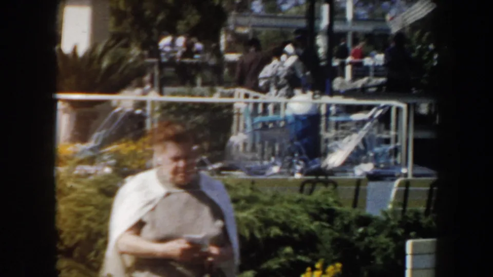1961 California frame with motion blur on a foreground woman, outdoor planting beds, railings, and visitors behind her.