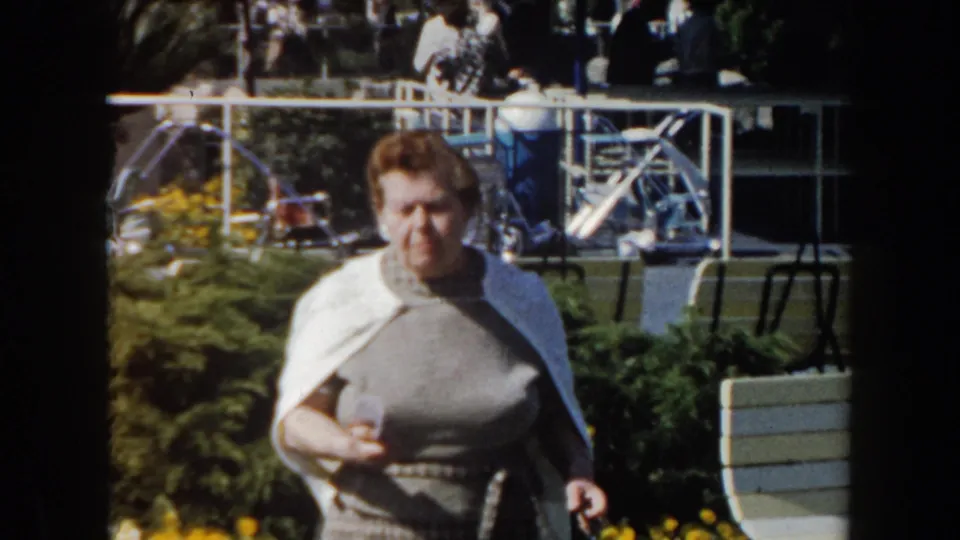 The woman approaches the camera in a California garden setting, with benches, railings, and people visible behind her.