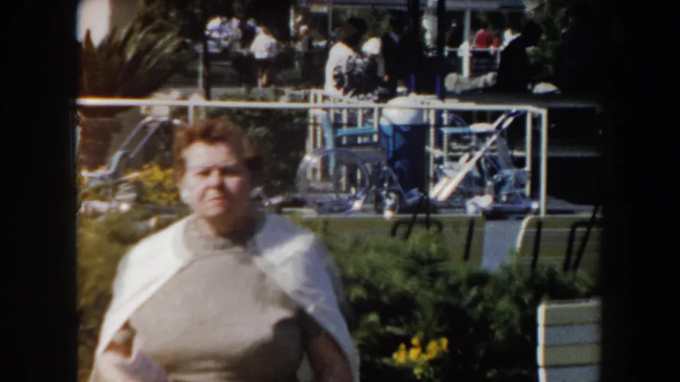 A woman in a gray sweater and white shawl is centered in the 1961 California garden scene, with flowers and recreation structures behind her.