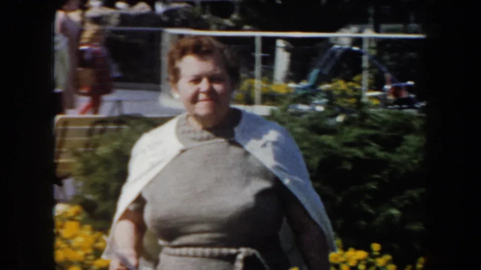 Near the end of the clip, the woman passes close to the camera in the garden, with yellow flowers and background movement visible.