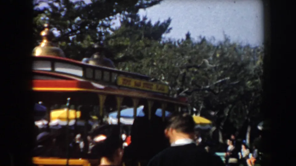 A red and yellow park trolley with partial Main Street Plaza signage passes through a tree-lined amusement park scene, with an adult visitor in foreground.