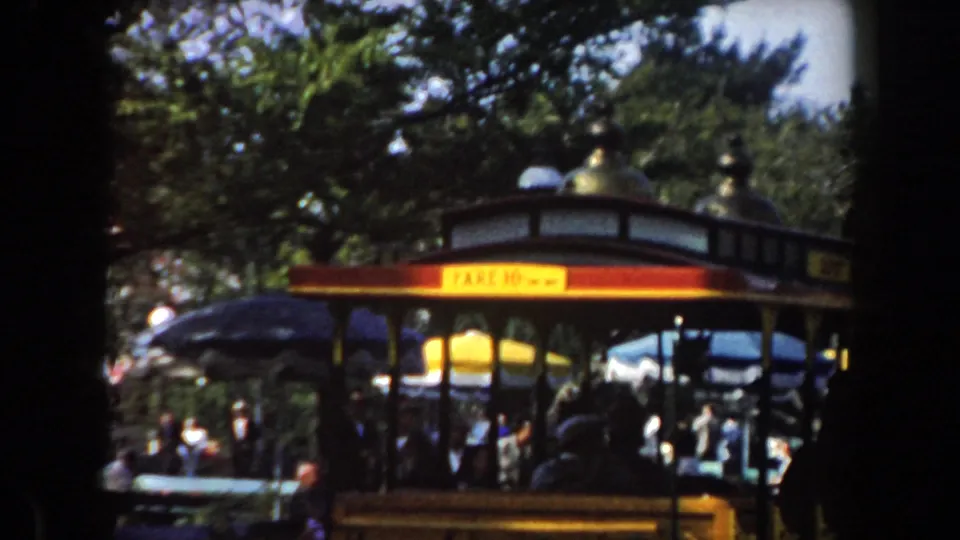 Red and yellow amusement park trolley carrying silhouetted riders, with benches, umbrellas, trees, and visitors in the background in 8mm film color.