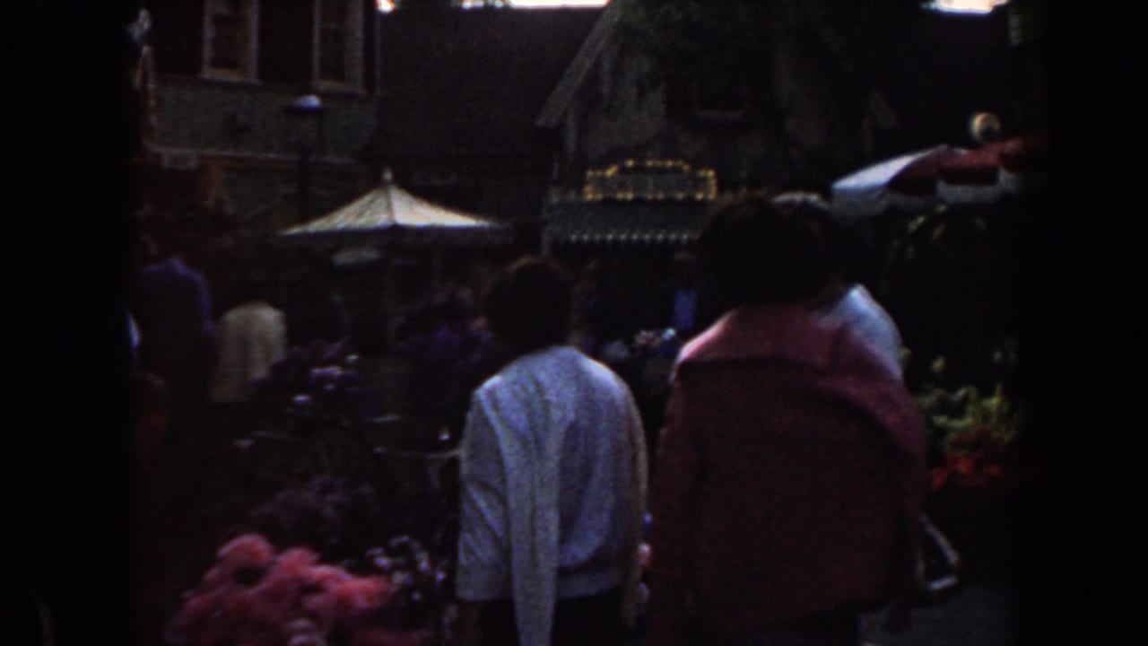 1961 California home-movie frame showing adults from behind near colorful flower stalls and a red and white canopy.