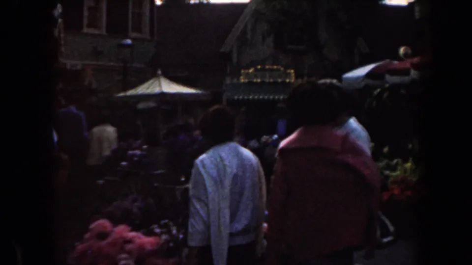 1961 California home-movie frame showing adults from behind near colorful flower stalls and a red and white canopy.