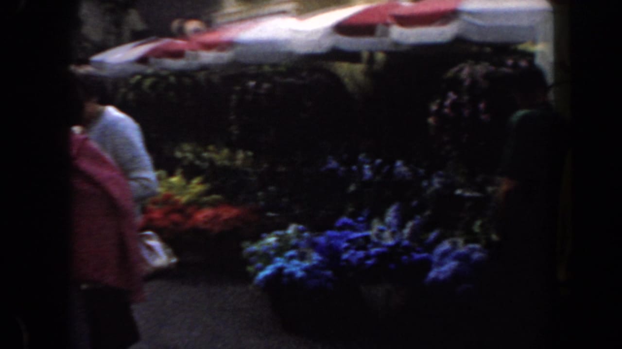 People pass flower stalls under a striped canopy in a dark fair walkway.