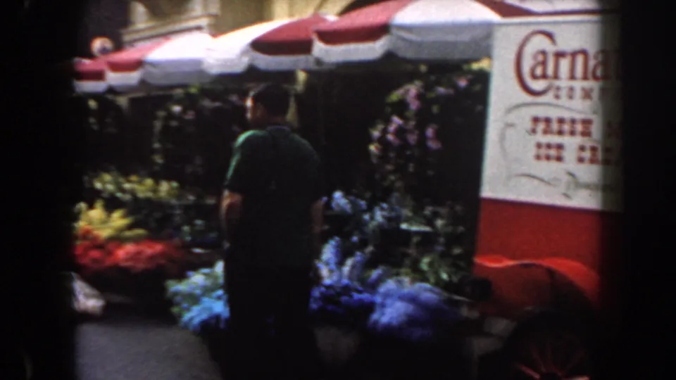 1961 California home-movie still with a visitor near flower stalls and a branded ice cream cart at a fair.