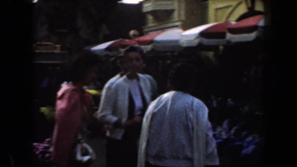 1961 California fair frame showing adults in a crowded floral market area with striped canopy shade.