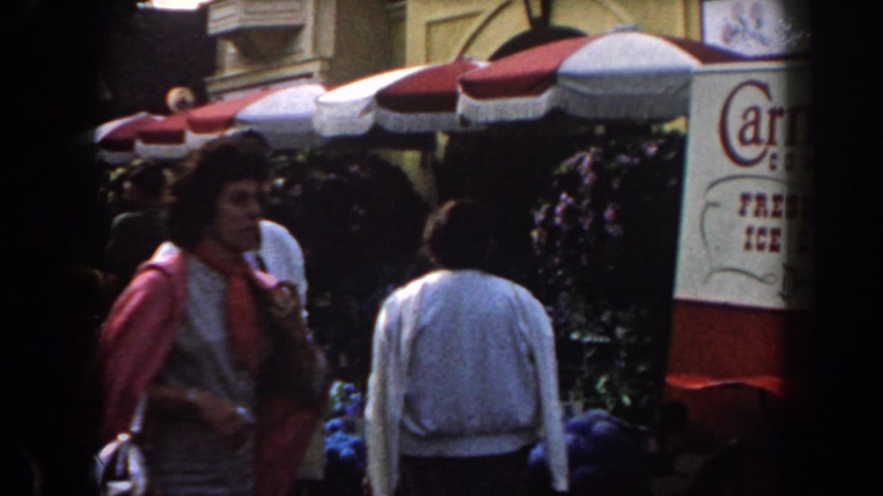 1961 California home-movie frame showing shoppers beside colorful flowers, striped umbrellas, and a partially visible market sign.