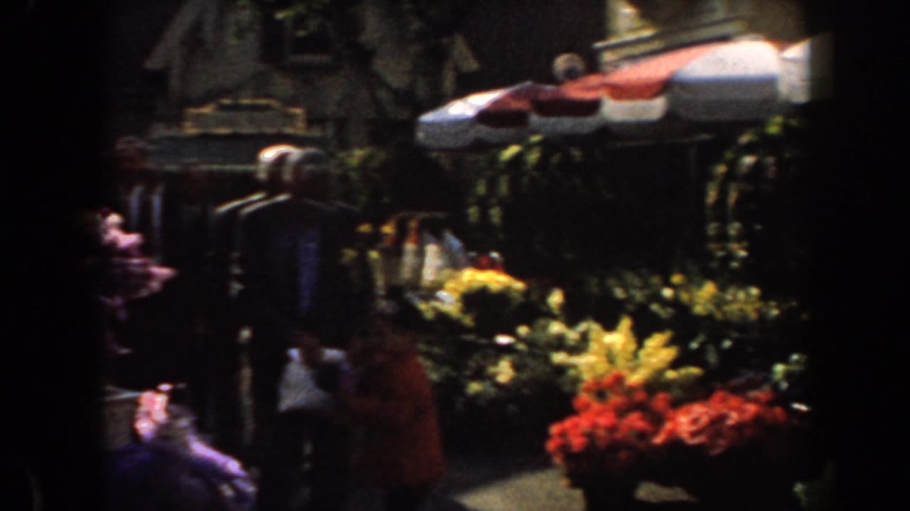 Flower displays and shoppers line a shaded outdoor market walkway.