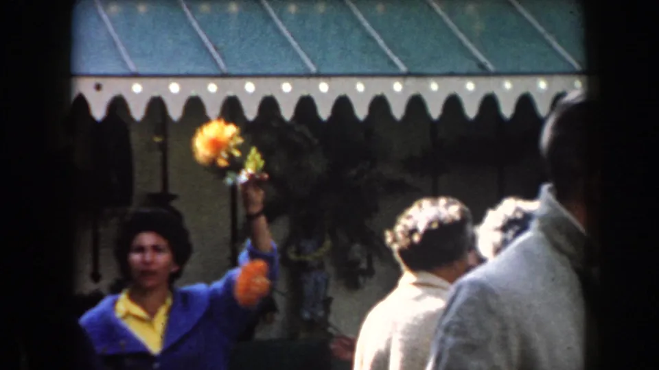 1961 California market frame showing a woman in a blue jacket lifting a yellow flower beside an awning and nearby shoppers.