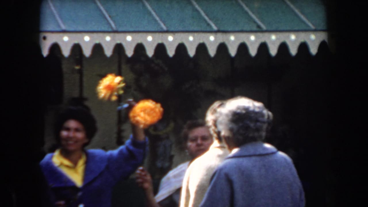 Woman in blue and yellow holds orange flowers beside a line of adults under a scalloped awning.