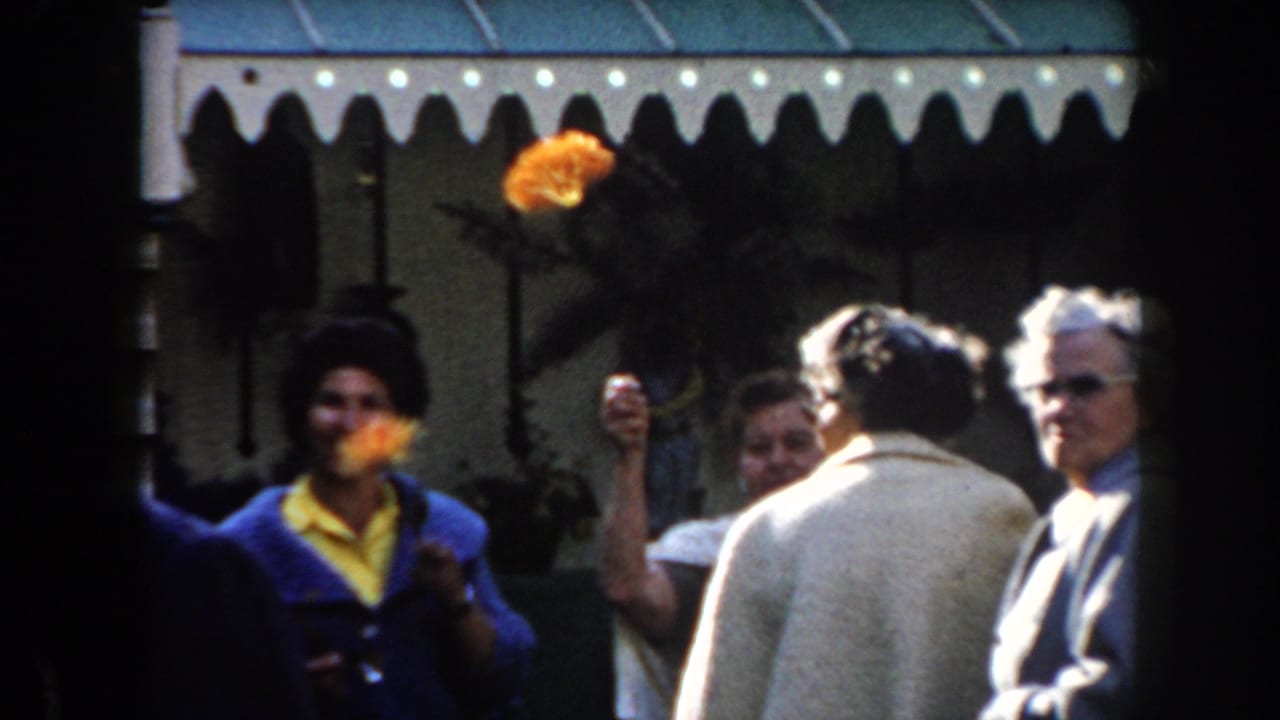 Women hold orange flowers as older adults stand and turn near a green-and-white awning.
