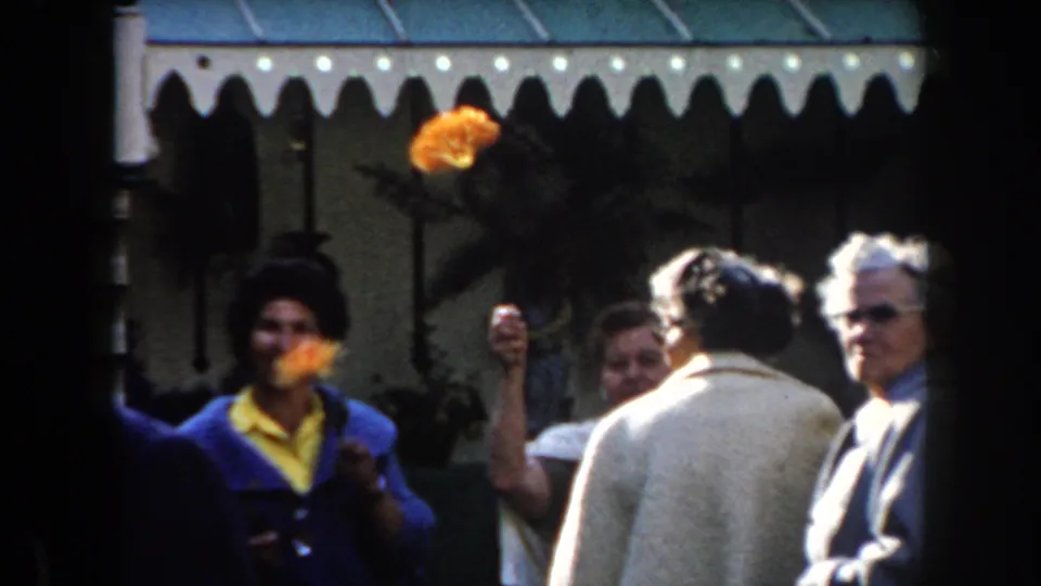 Women in blue and yellow hold orange flowers while older adults in light jackets gather outdoors in the 1961 California home-movie scene.
