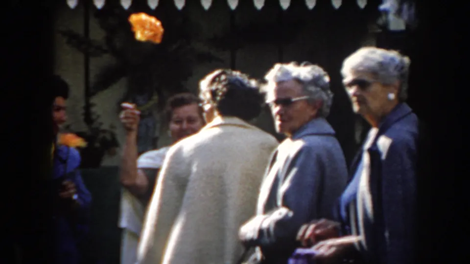 A group of adults, including people in sunglasses and light coats, stands near raised orange flowers in a 1961 California outdoor home-movie shot.