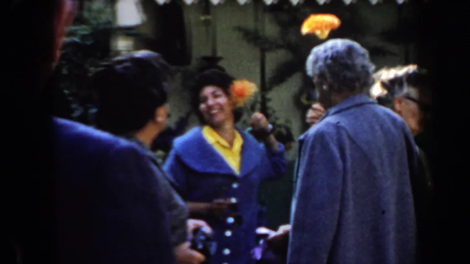 A smiling woman in a blue jacket and yellow shirt holds orange flowers toward adults in a sunny 1961 California home-movie gathering.