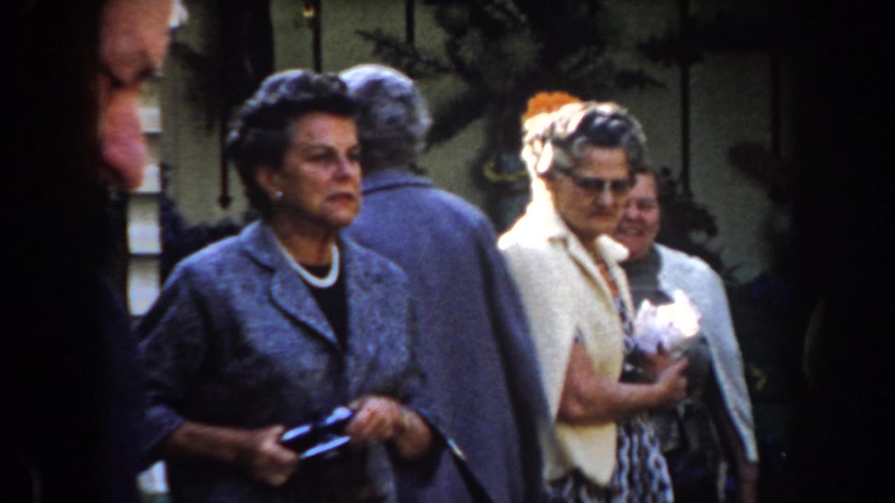 Older women stand outdoors in coats and hats, with one holding a dark handbag or camera case.