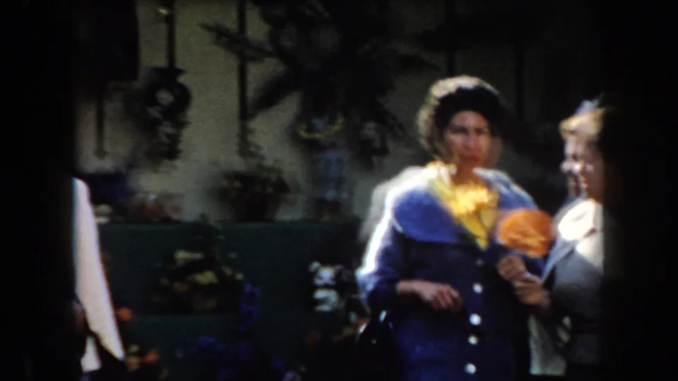 Two older women, one in a blue jacket and yellow blouse, speak outdoors while holding orange flowers in a garden setting.
