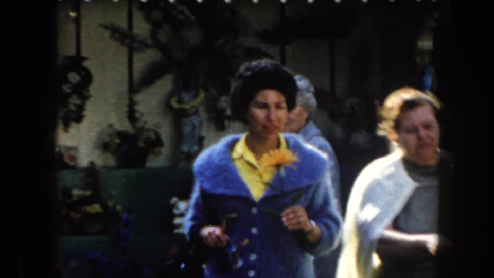 Older women in California archival footage stand in strong sunlight, one holding an orange flower and sunglasses near garden decor.