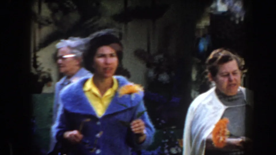 In a 1961 California garden scene, two adult women walk past greenery while one wears a blue jacket over a yellow shirt.