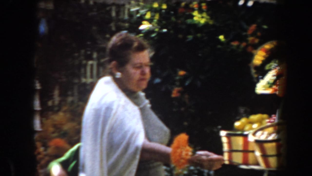 Woman in a white shawl holding orange flowers beside baskets of produce.