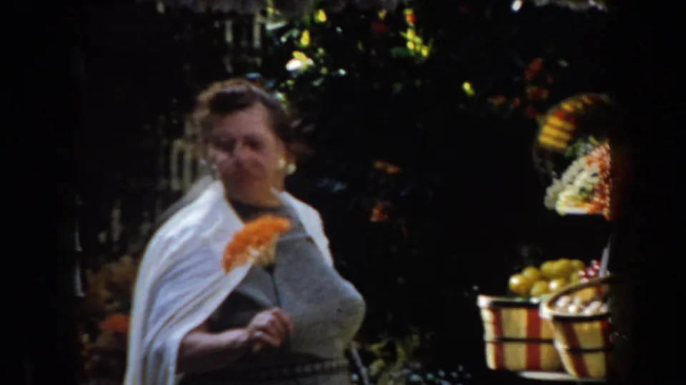 The woman in a white shawl holds orange flowers while standing beside foliage and a produce display in California footage from 1961.