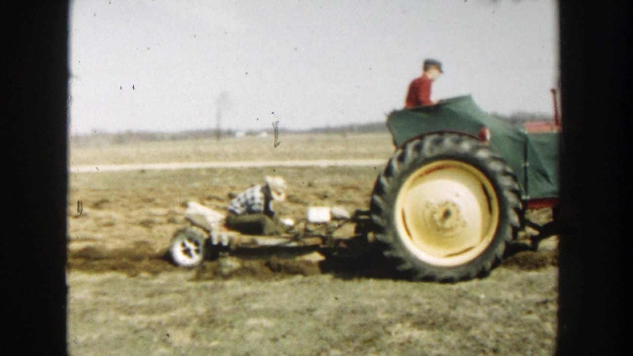 1954 Wisconsin Dells farm scene with a green tractor, seated operator, and a man in plaid tending a trailing implement in a dry field.
