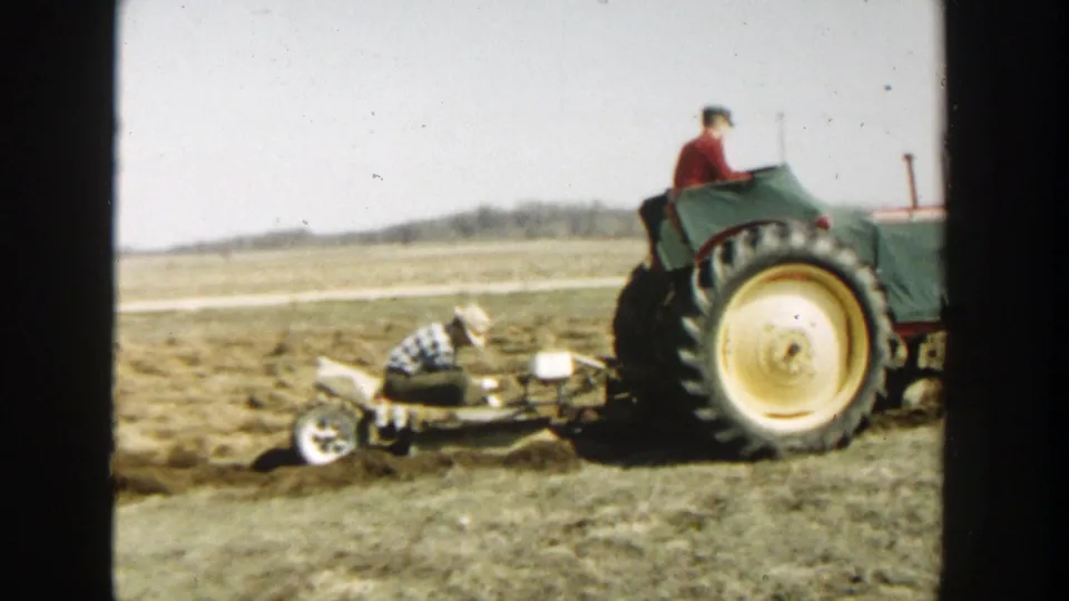 The 1954 Wisconsin Dells home movie shows a plaid-shirted worker beside the implement as the green tractor moves through dry soil.