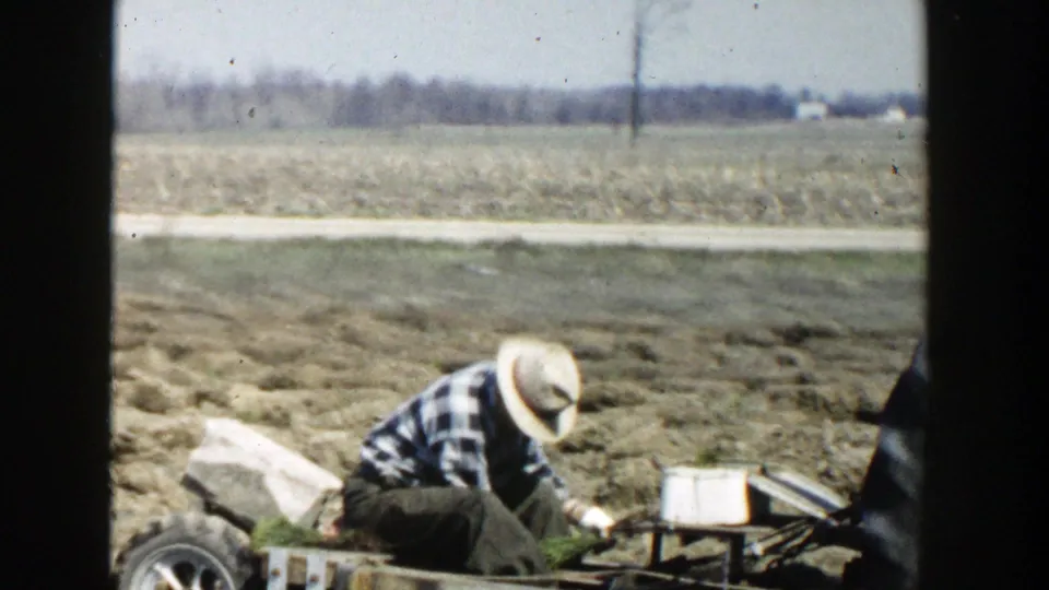 1954 Wisconsin Dells home-movie frame showing a farmer in a plaid shirt and straw hat tending equipment in a rural field.