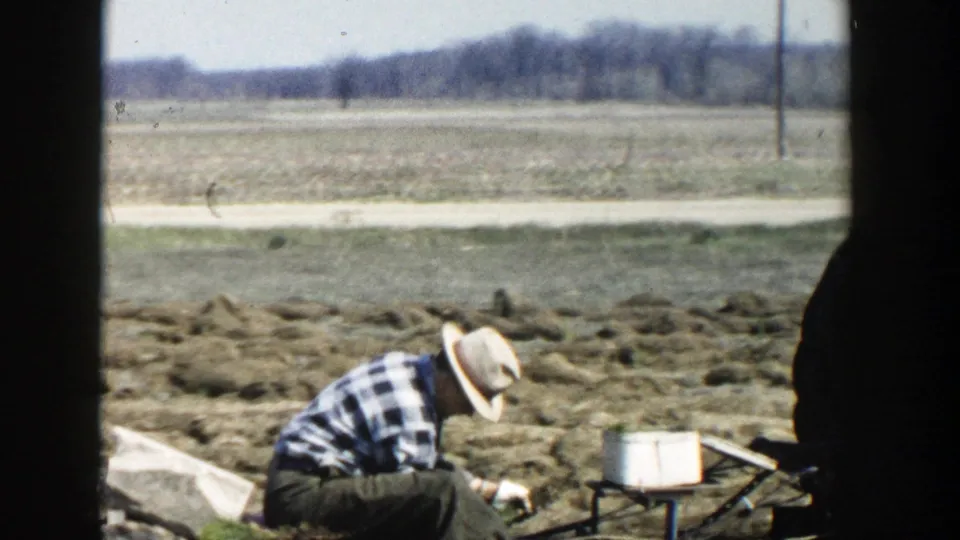 A farmer in work clothes tends a tractor-mounted attachment in an open Wisconsin Dells field, seen through grainy 8mm color film.