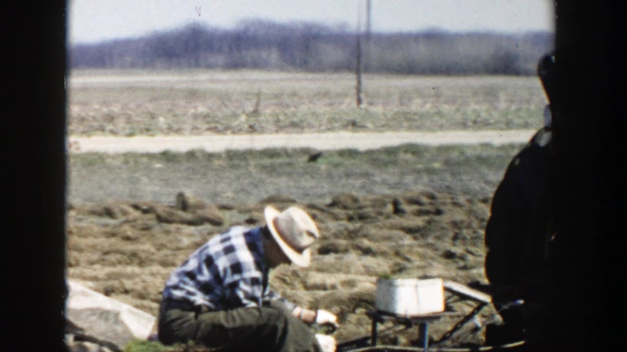 In 1954 Wisconsin Dells footage, a farmer in a plaid shirt and straw hat works low to the ground near a white pail in a tilled field.