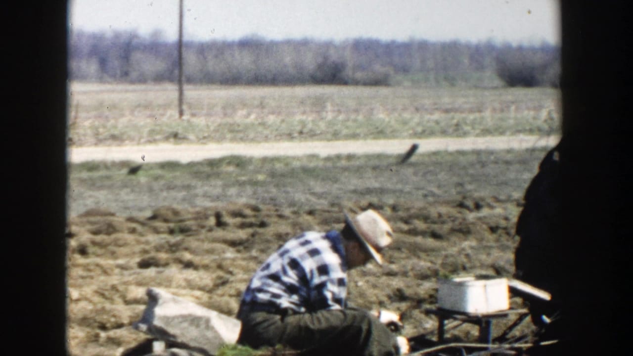 Farmer crouches in a tilled field as a white container sits on a small frame nearby.