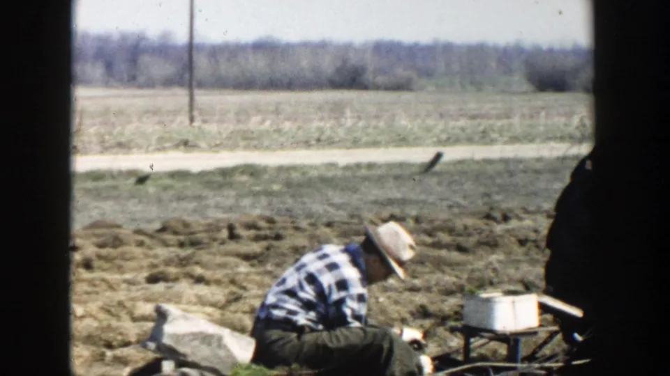 The farmer remains crouched near the soil in the Wisconsin Dells farm scene, with open land and a distant treeline behind him.