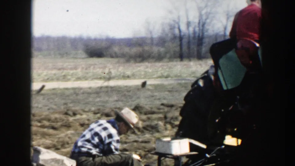 A rider on a tractor moves into the frame beside the crouching farmer, with tilled soil and open Wisconsin countryside visible beyond.