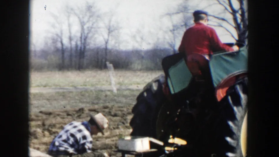 The tractor rider sits above the field equipment while the crouching farmer remains at work in the 1954 Wisconsin Dells farm footage.