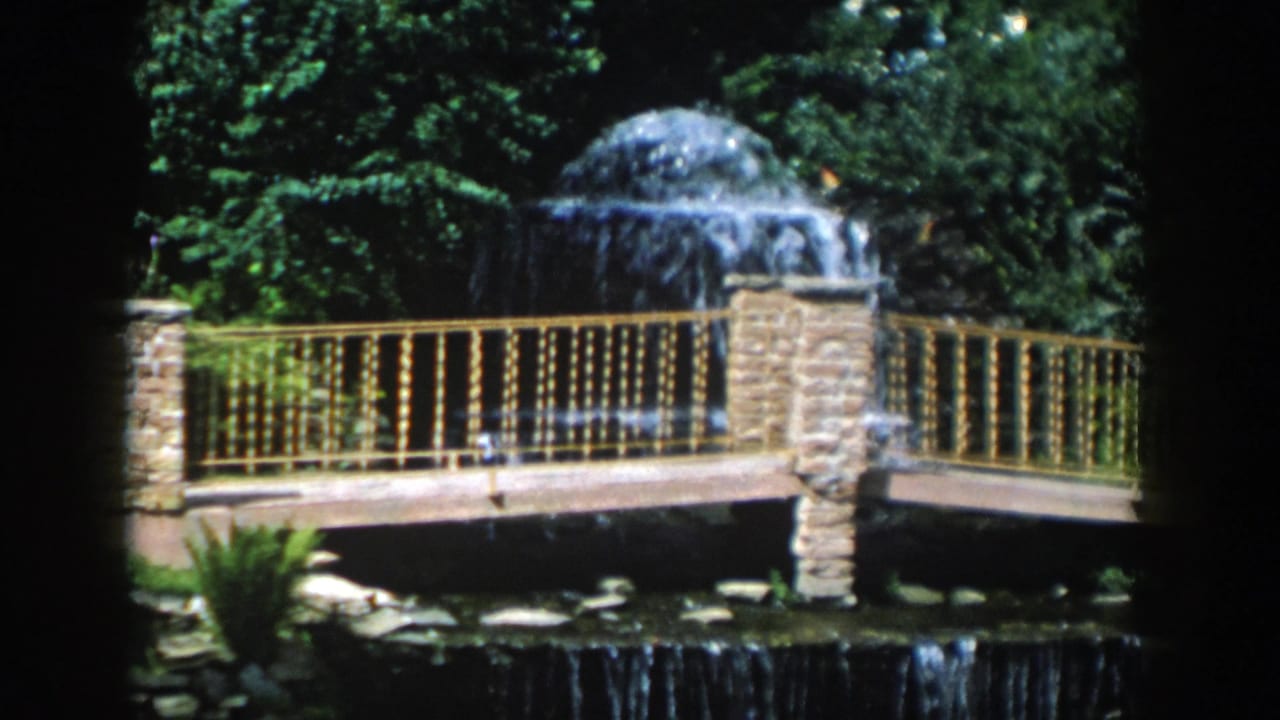 Stone fountain and bridge railing above a small waterfall surrounded by greenery.