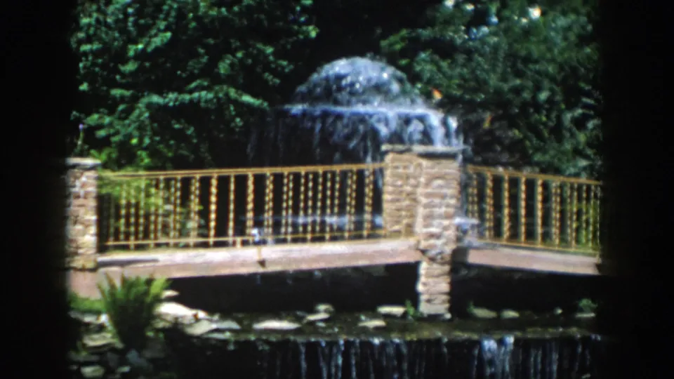 1954 Wisconsin Dells 8mm frame showing a stone fountain, ornate railing, bridge, pond edge, and surrounding trees.