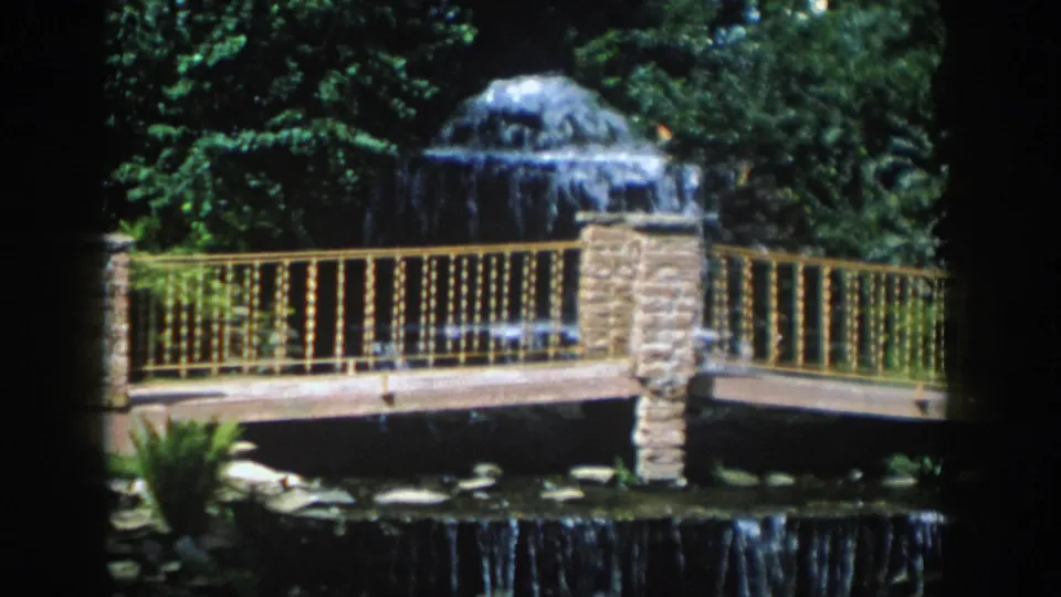 1954 Wisconsin Dells 8mm frame with soft-focus garden scenery, a fountain, railing, bridge, and shaded water below.