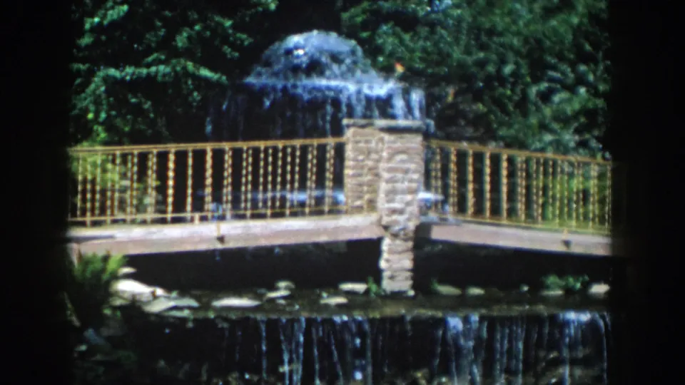 1954 Wisconsin Dells frame showing a landscaped fountain scene with a bridge, railing, stonework, foliage, and lower cascade.
