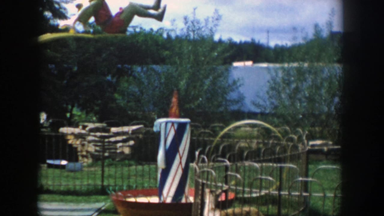 1954 Wisconsin Dells home-movie frame showing a clown-topped yellow ride, striped post, fenced yard, trees, and distant water.