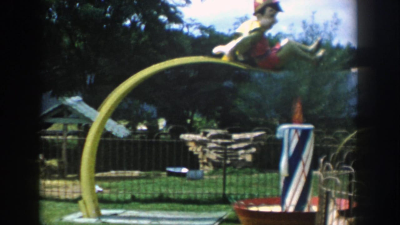 1954 Wisconsin Dells frame with a yellow arched ride, clown figure, striped post, low fencing, stone wall, and trees behind it.
